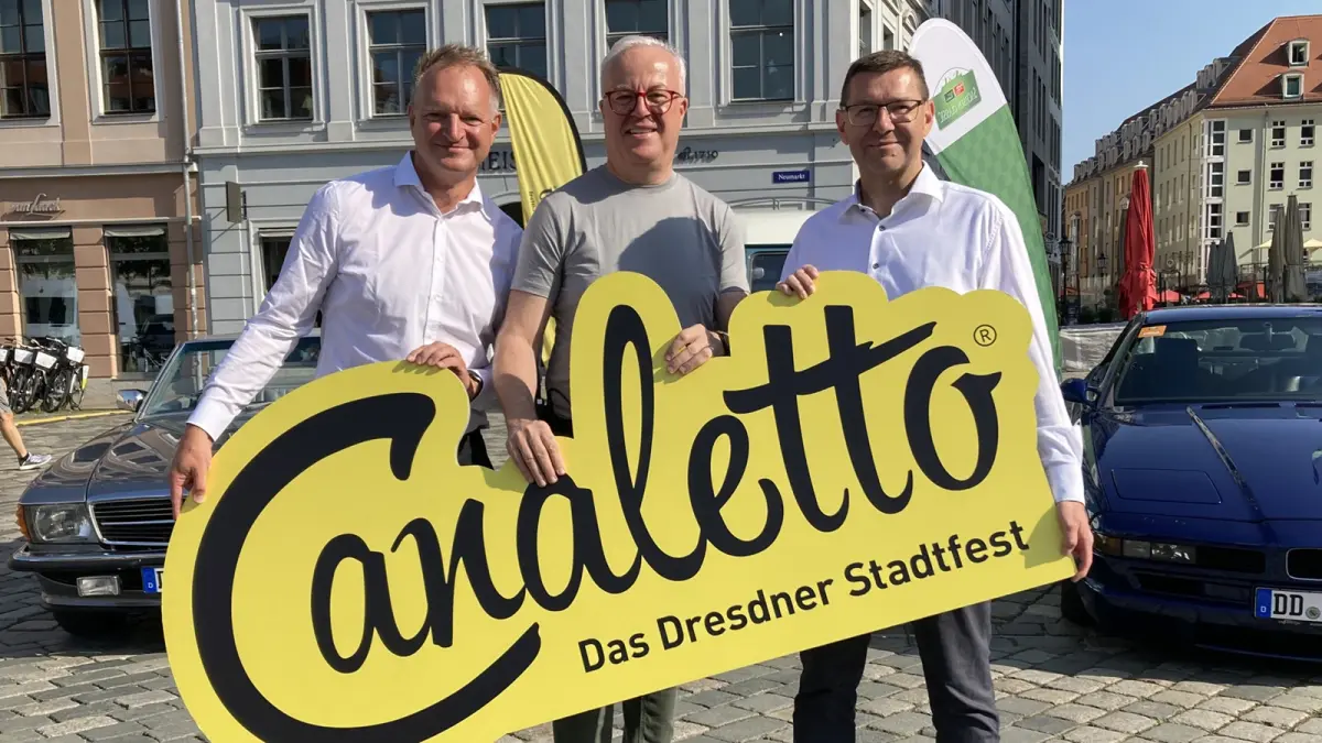 Drei Männer halten ein gelbes Schild mit der Aufschrift Canaletto Das Dresdner Stadtfest. Im Hintergrund Autos und Häuser.