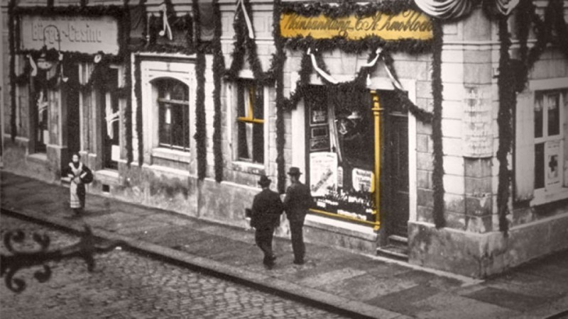 Zwei Männer gehen an geschmückten Ladenfronten einer gepflasterten Straße entlang. Eine Frau steht weiter entfernt.