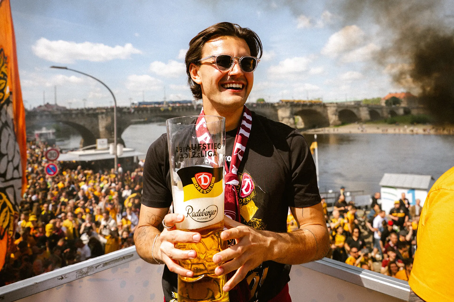 Ein junger Mann hält ein großes Bierglas und steht vor einer Menschenmenge an einem sonnigen Tag. Er trägt eine Sonnenbrille und einen Schal.