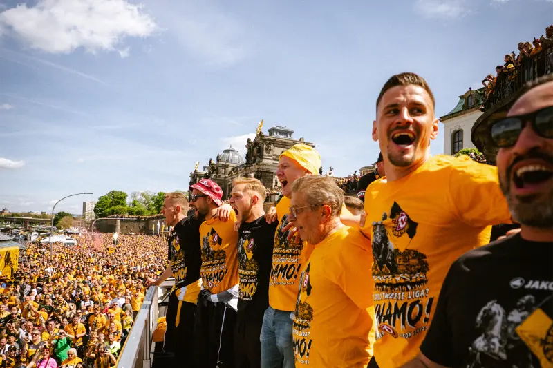 Eine Gruppe von Menschen in gelben T-Shirts jubelt auf einer Bühne vor einer Menge.
