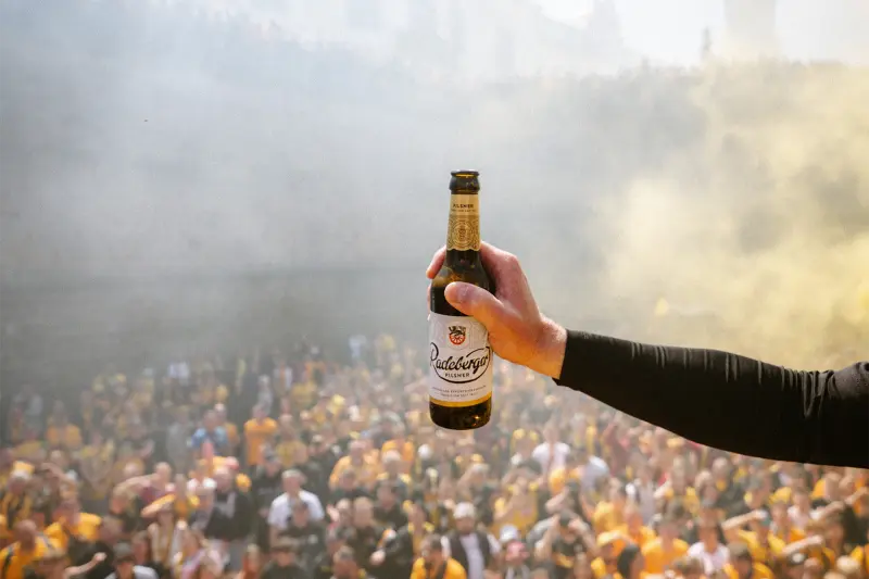 Eine Hand hält eine Flasche Bier vor einer Menschenmenge im Freien.