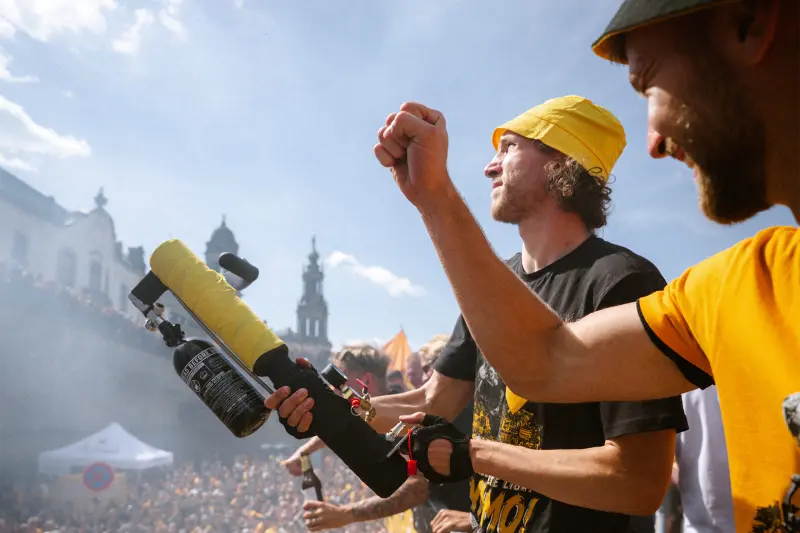 Zwei junge Männer in gelben Hüten freuen sich bei einem sonnigen Festival. Einer hält eine Konfettikanone.