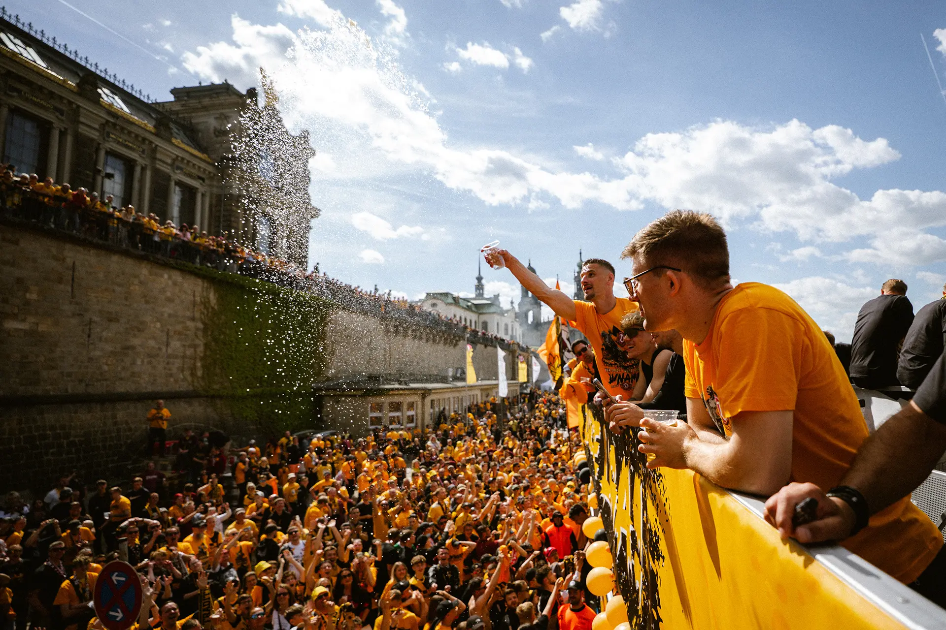 Mehrere Personen in gelben Shirts feiern auf einer Straße, Wasser wird verspritzt.