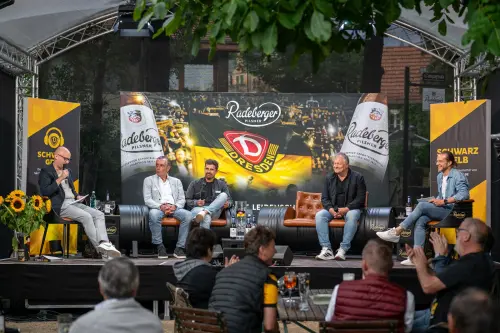 Fünf Männer sitzen auf einer Bühne vor großem Banner von Dynamo Dresden und Radeberger. Im Vordergrund sitzen Zuschauer.