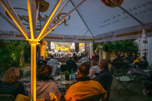 Zuschauer sitzen unter Schirmen vor einer Bühne mit fünf Männern. Im Hintergrund Banner mit Dynamo Dresden und Radeberger Werbung.