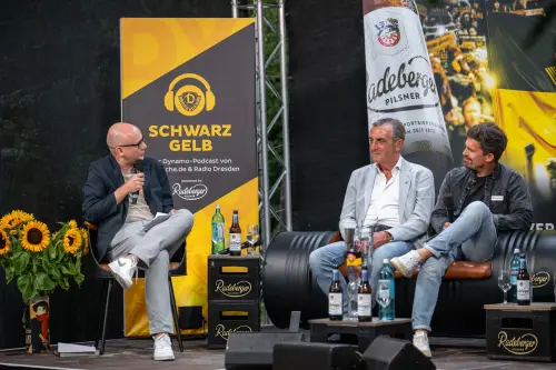 Drei Männer sitzen auf einer Bühne vor Banner von Dynamo Dresden und Radeberger. Links ein Plakat mit Werbung für Schwarz-Gelb Podcast.