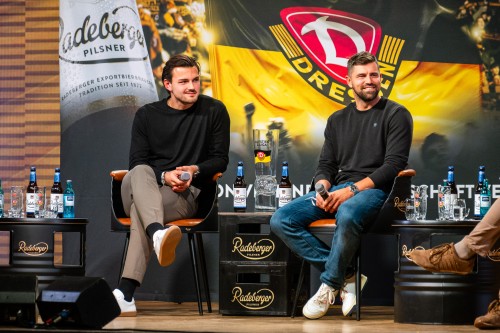 ZWEI Männer sitzen auf einer Bühne vor großem Banner von Dynamo Dresden und Radeberger.