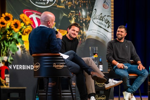 Drei Männer sitzen auf einer Bühne vor großem Banner von Dynamo Dresden und Radeberger.In einer Interviewsituation.