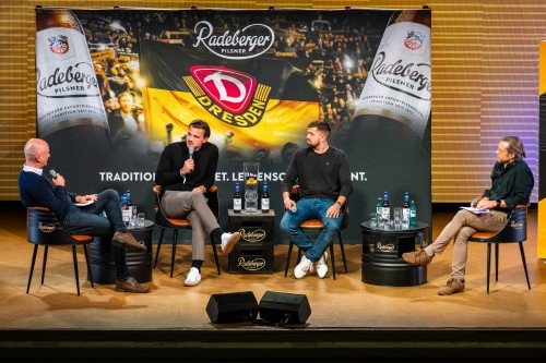Vier Männer sitzen auf einer Bühne vor großem Banner von Dynamo Dresden und Radeberger. In einer Interviewsituation.