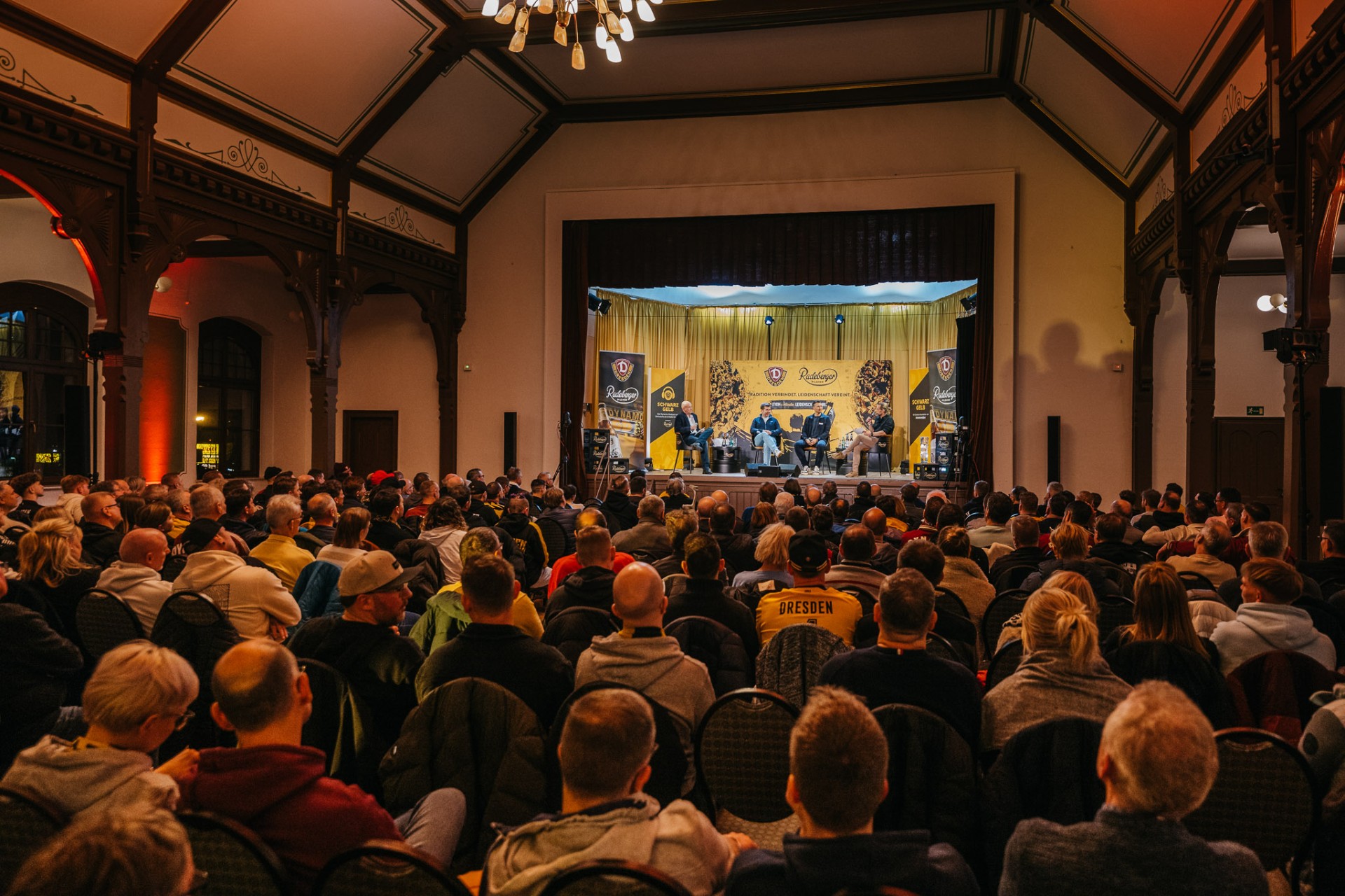 Ein Saal ist voll besetzt mit Zuschauern, auf der Bühne befinden sich mehrere Personen an einem Tisch in einer Interview Situation.