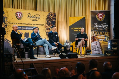 Vier Männer sitzen auf einer Bühne vor einem großem Banner von Dynamo Dresden und Radeberger. Sie sprechen miteinander.