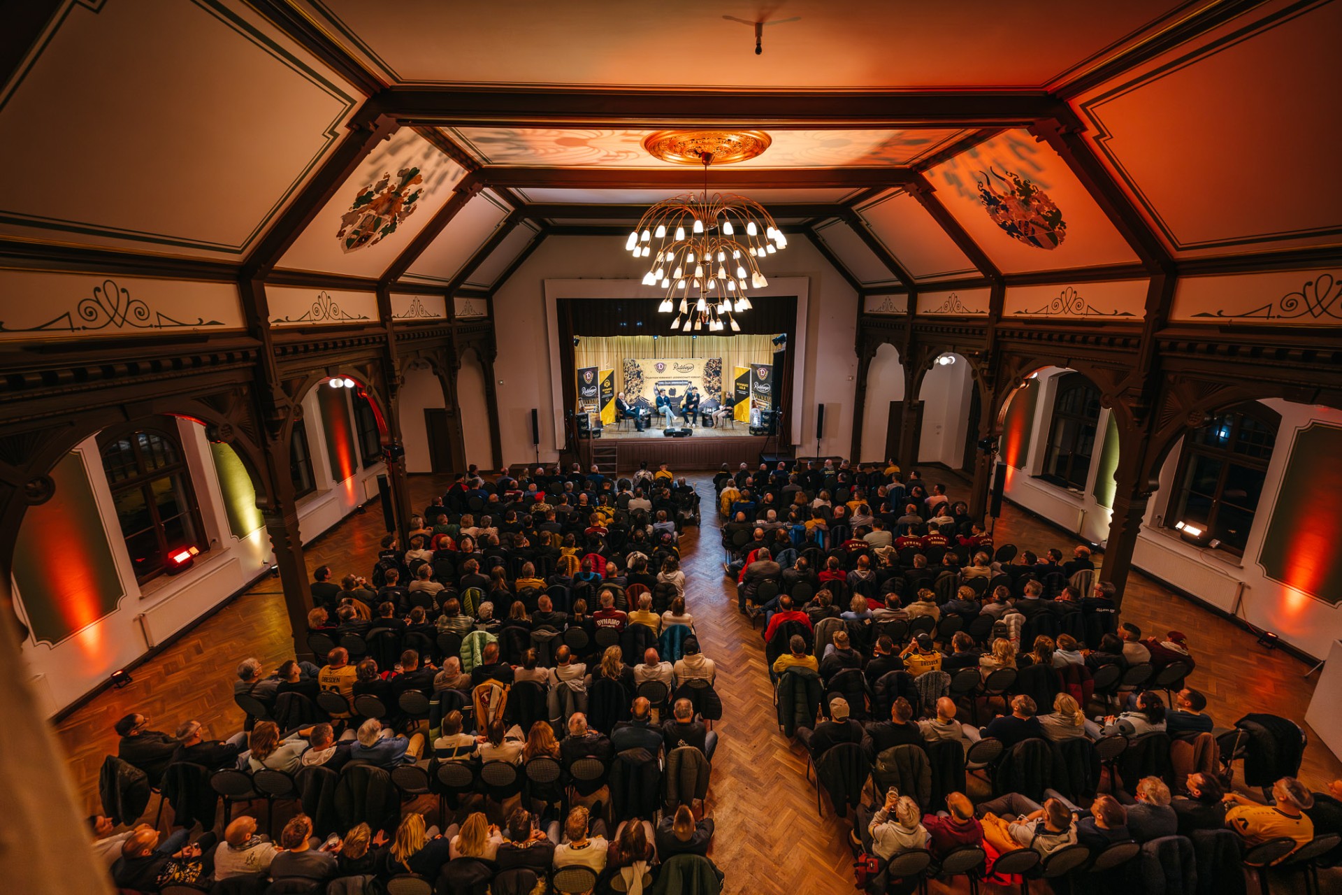 Ansicht von oben in einen Saal der voll besetzt mit Zuschauern ist, auf der Bühne befinden sich mehrere Personen an einem Tisch in einer Interview Situation.