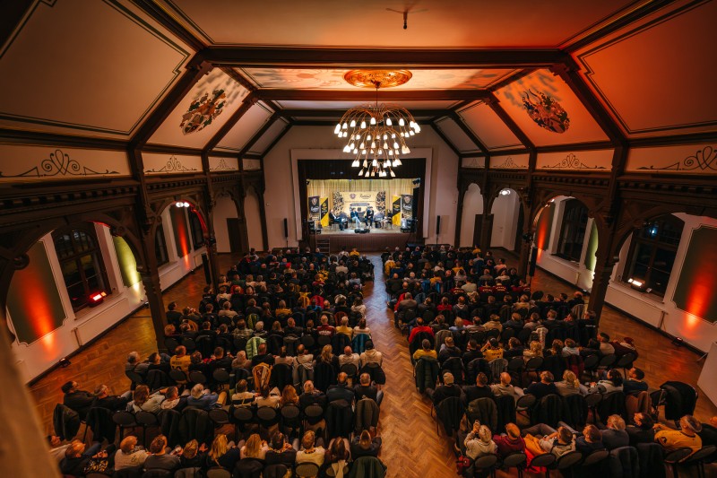 Ansicht von oben in einen Saal der voll besetzt mit Zuschauern ist, auf der Bühne befinden sich mehrere Personen an einem Tisch in einer Interview Situation.
