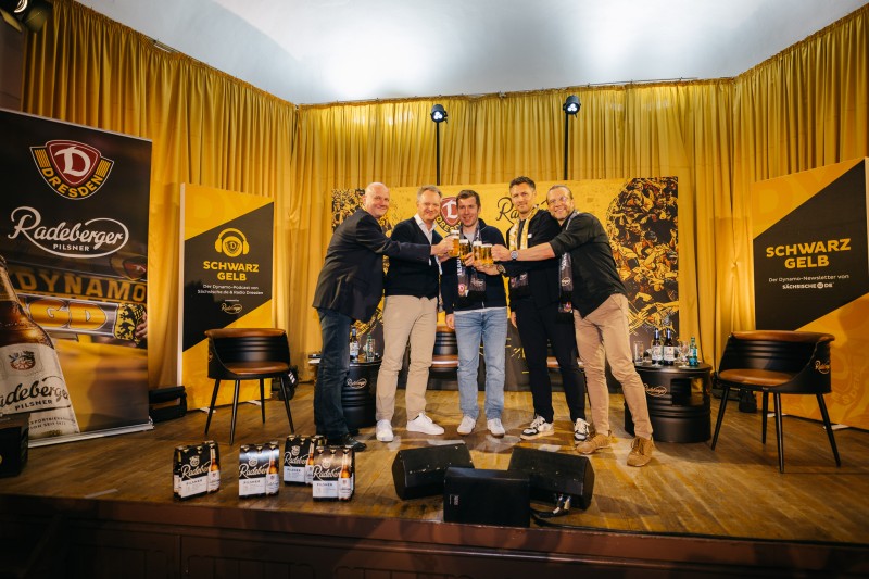 Fünfi Männer stehen auf einer Bühne vor einem großem Banner von Dynamo Dresden und Radeberger. SIe stoßen mit Bier an.