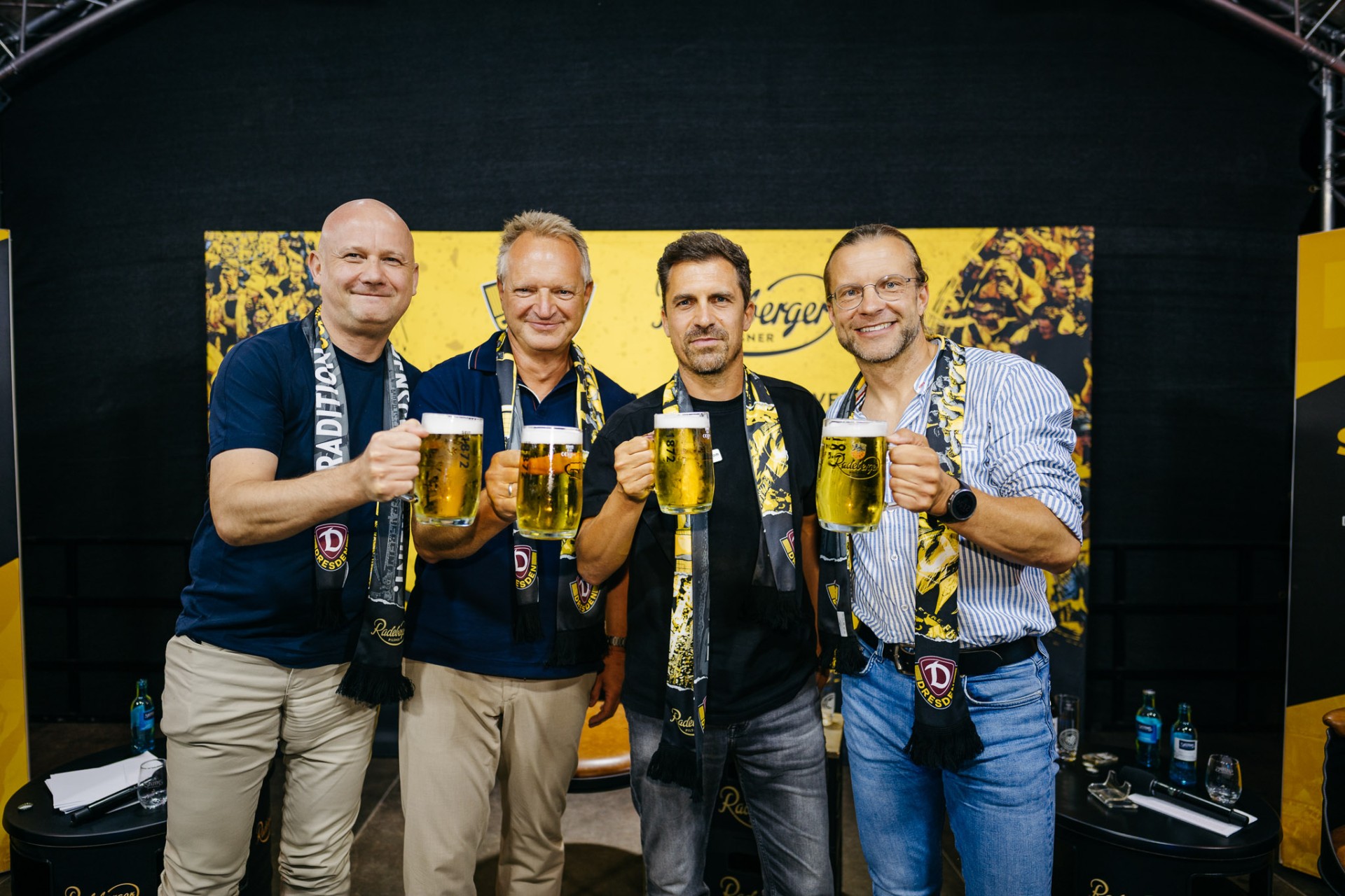 Gruppe von vier Männern mit Radeberger Bierkrügen in der Hand und Dynamo Dresden Schals um den Hals.