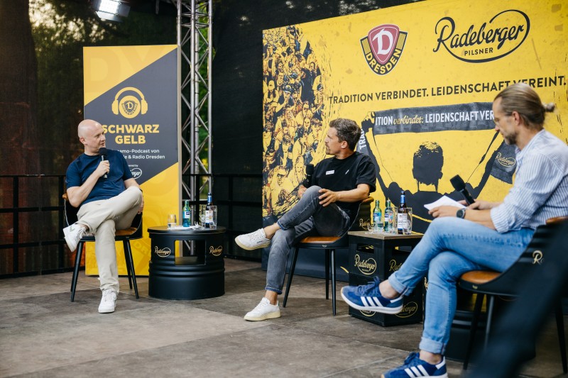Drei Männer sitzen auf einer Bühne und diskutieren. Im Hintergrund sind Banner von Dynamo Dresden und Radeberger Pilsner.