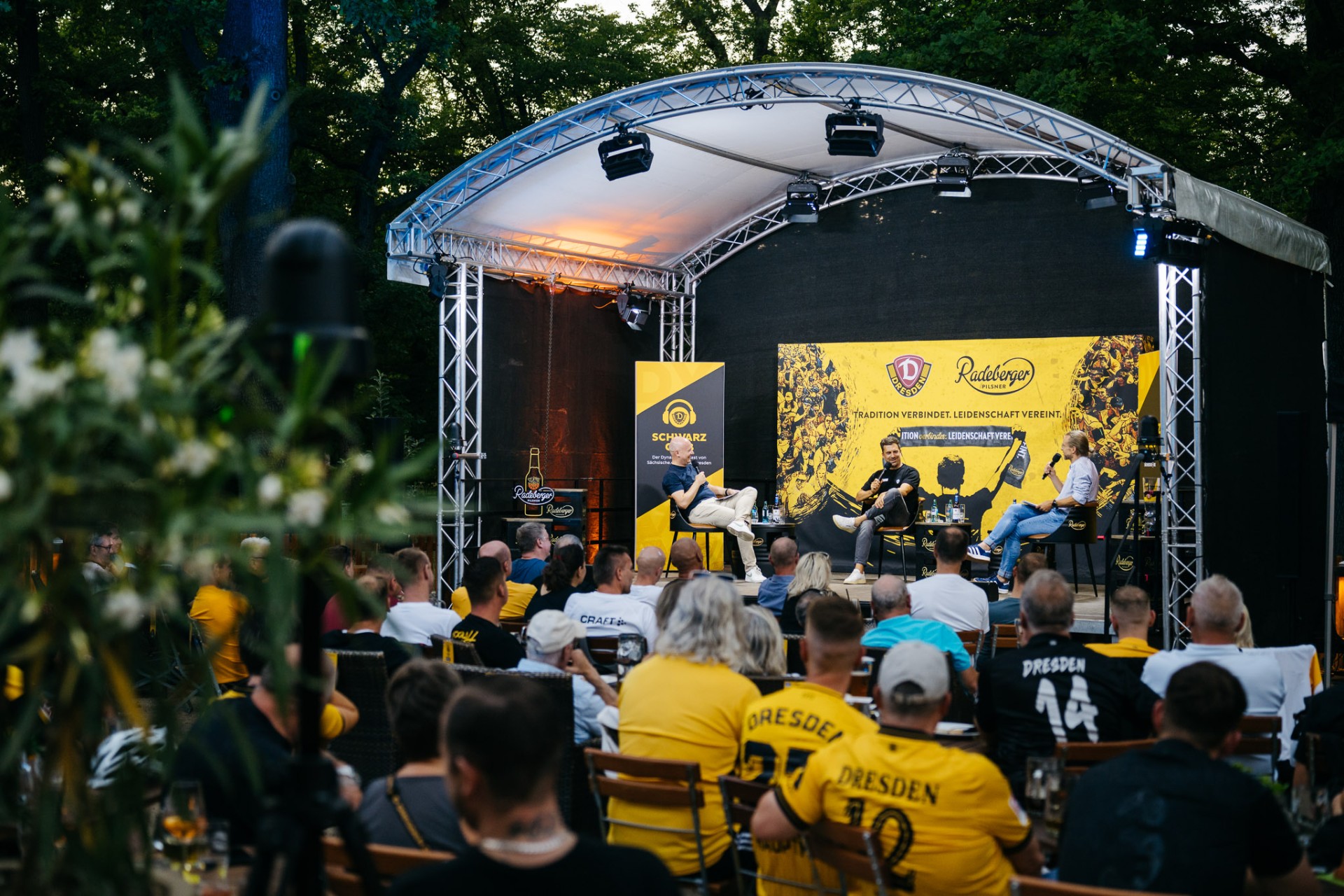 Zuschauer verfolgen eine Podiumsdiskussion mit drei Personen hinter Dynamo Dresden und Radeberger Pilsner Werbebannern.