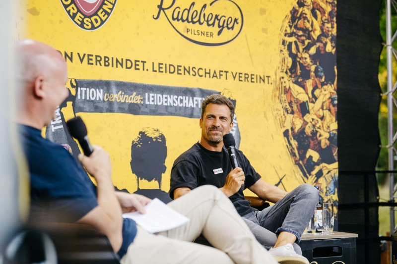 Zwei Männer sitzen auf einer Bühne und halten Mikrofone. Im Hintergrund Werbenanner von Radeberger und Dynamo Dresden.