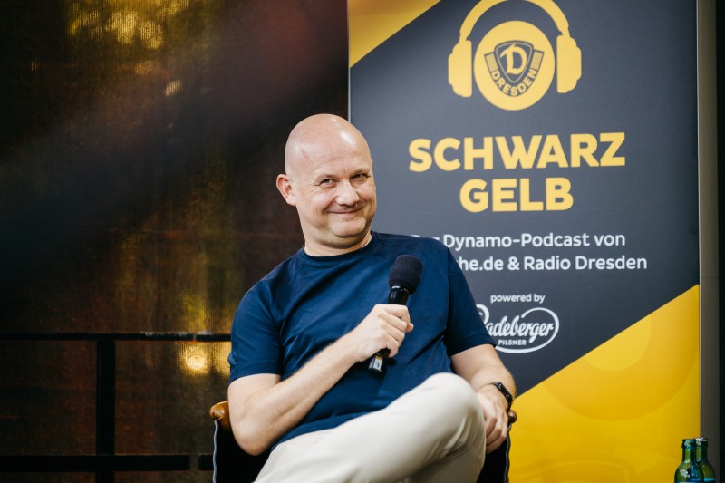 Lächelnder Mann sitzt vor einem Banner mit Dynamo Dresden und Radeberger Werbung. Er trägt ein Mikrofon in der rechten Hand.