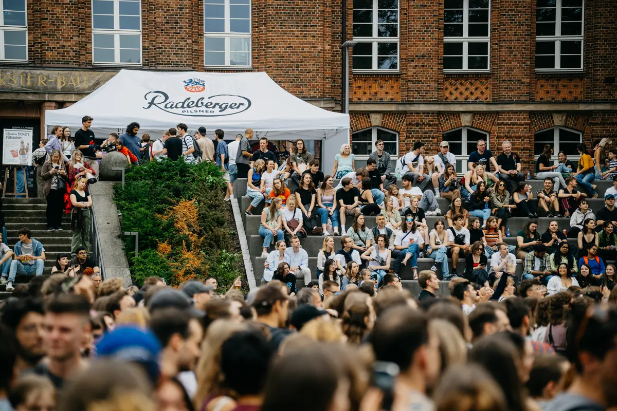 Viele Menschen sitzen und stehen vor einem Ziegelgebäude bei einer Veranstaltung.