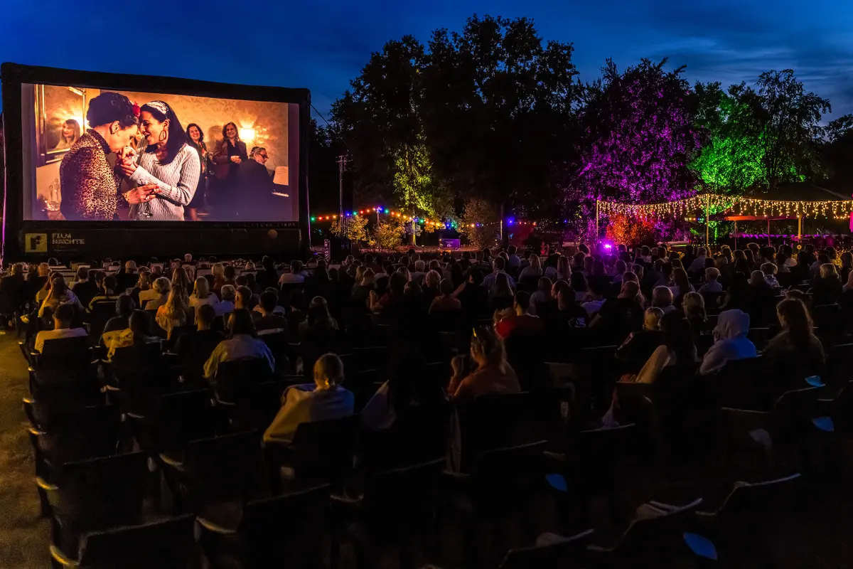 Eine Menschenmenge betrachtet bei Nacht einen Film in einem Open-Air-Kino mit bunt beleuchteten Bäumen.