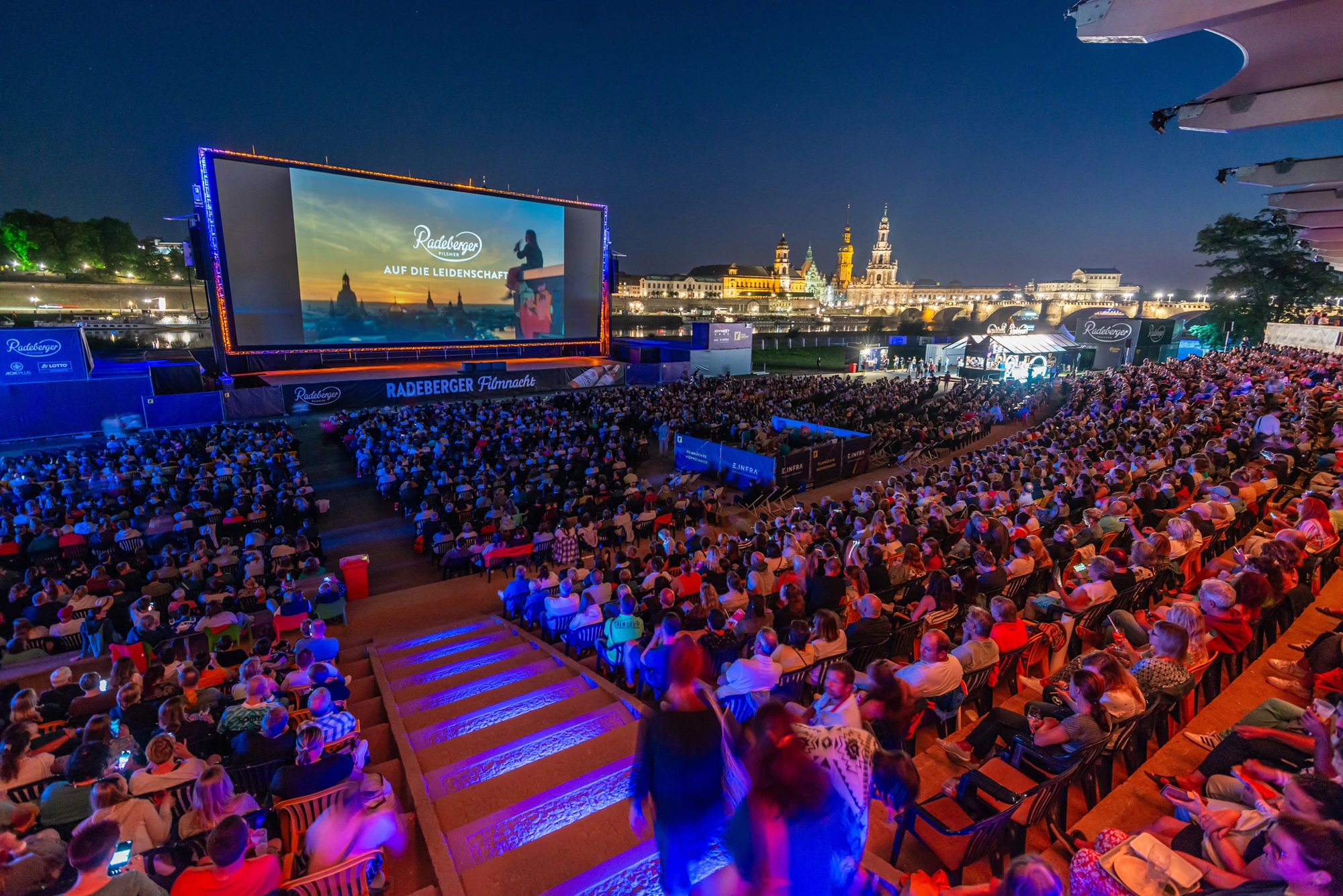 Viele Menschen sitzen bei Nacht im Freilichtkino vor einer großen Leinwand mit historischer Stadtsilhouette im Hintergrund.