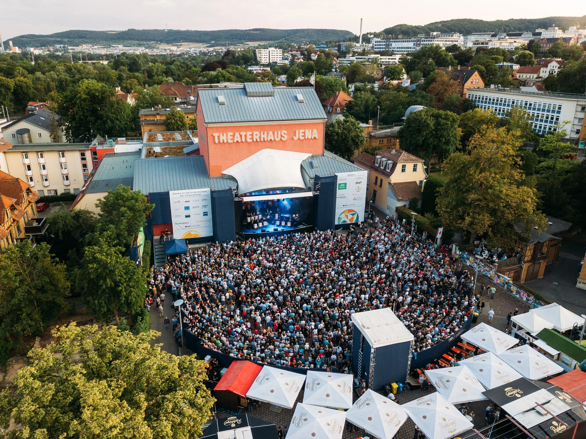 Viele Menschen stehen bei einem Open-Air-Event vor einer Bühne in einer städtischen Umgebung.