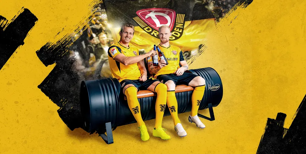 Zwei Fußballspieler in gelben Trikots sitzen mit Radeberger Bierflaschen vor einer großen Dynamo Dresden Fahne.