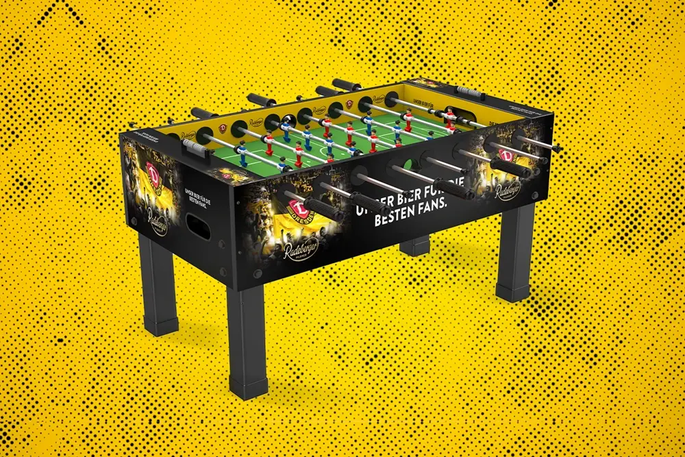 Schwarzer Tischkicker mit farbigen Spielfiguren im Dynamo-Dresden-Design. Gelber Hintergrund mit schwarzem Punktmuster.