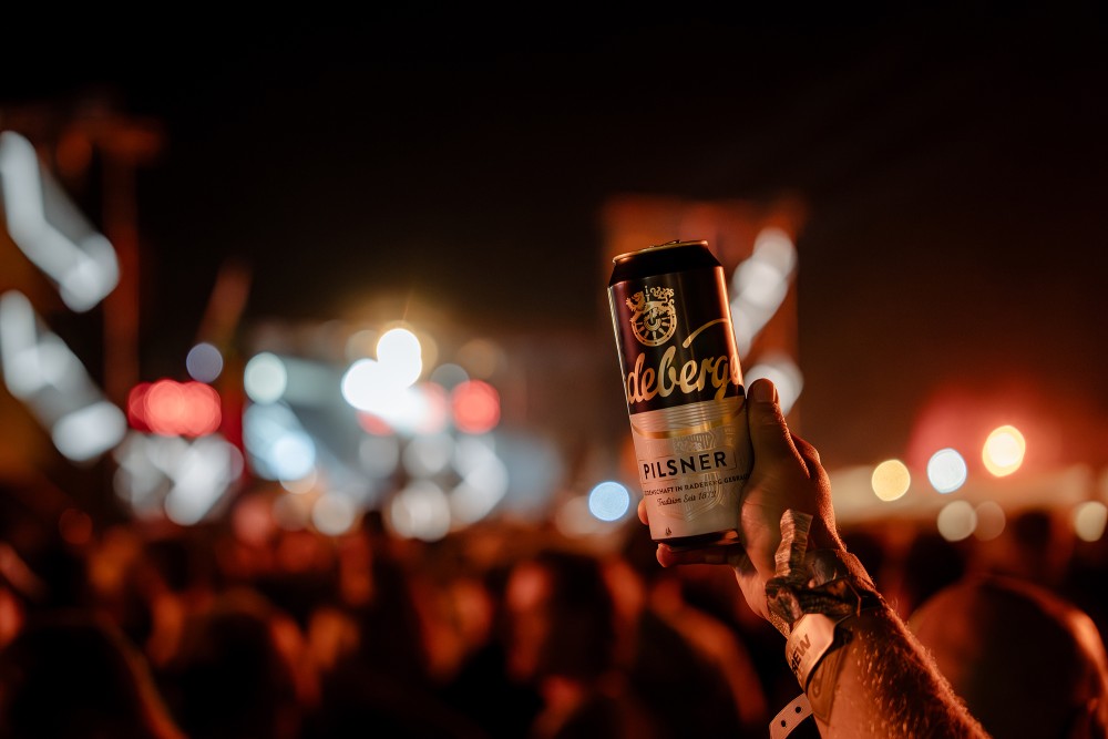 Eine große Menschenmenge feiert bei einem Open-Air-Konzert vor einer beleuchteten Bühne, eine Dose Radeberger im Vordergrund.