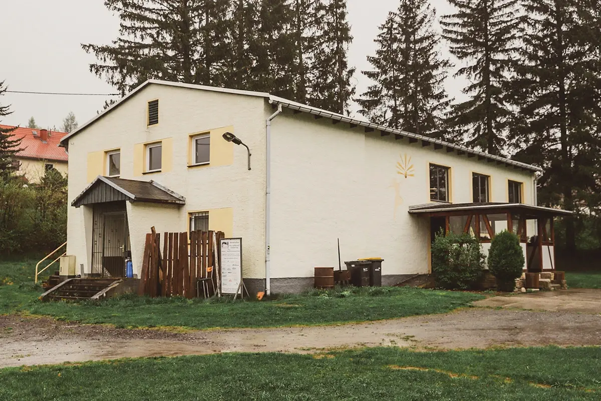 Ein helles, zweistöckiges Gebäude steht neben Bäumen, mit einer bedeckten Veranda und einer kleinen Rampe.
