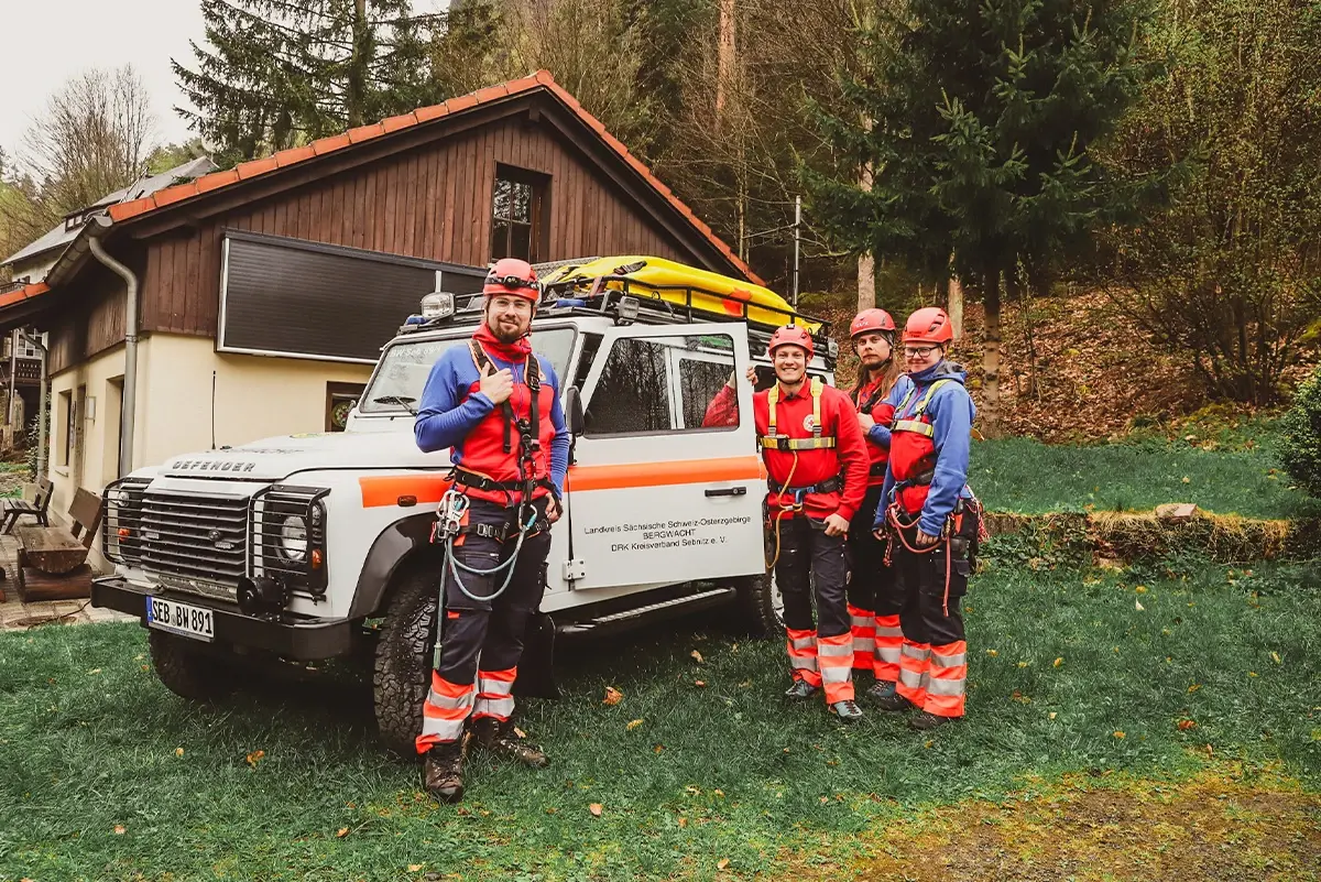 Fünf Personen in Schutzkleidung stehen neben einem Geländefahrzeug im Wald.