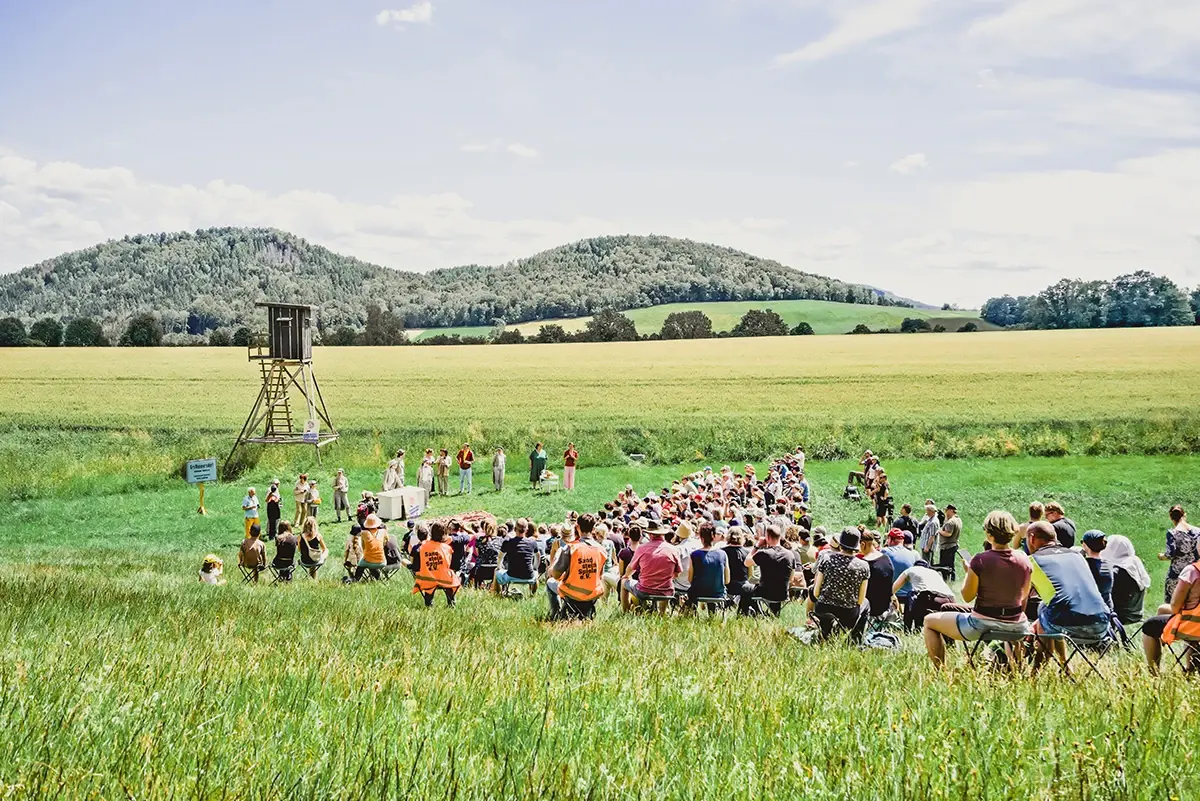 Eine große Menschenmenge sitzt auf einem Feld und lauscht einer Vorführung. Im Hintergrund sind grüne Hügel und ein Hochsitz.