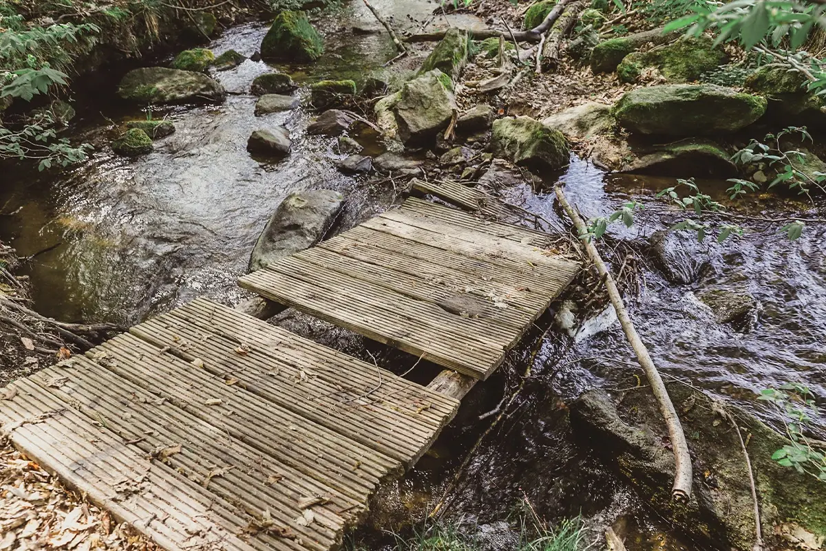 Ein hölzerner Steg überquert einen kleinen, felsigen Bach in einem bewaldeten Gebiet.