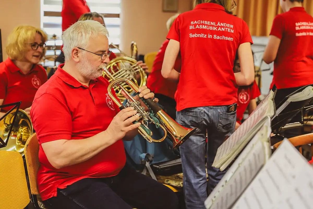 Ein Mann spielt Trompete in einer Gruppe mit roten Hemden. Sie proben in einem Raum.