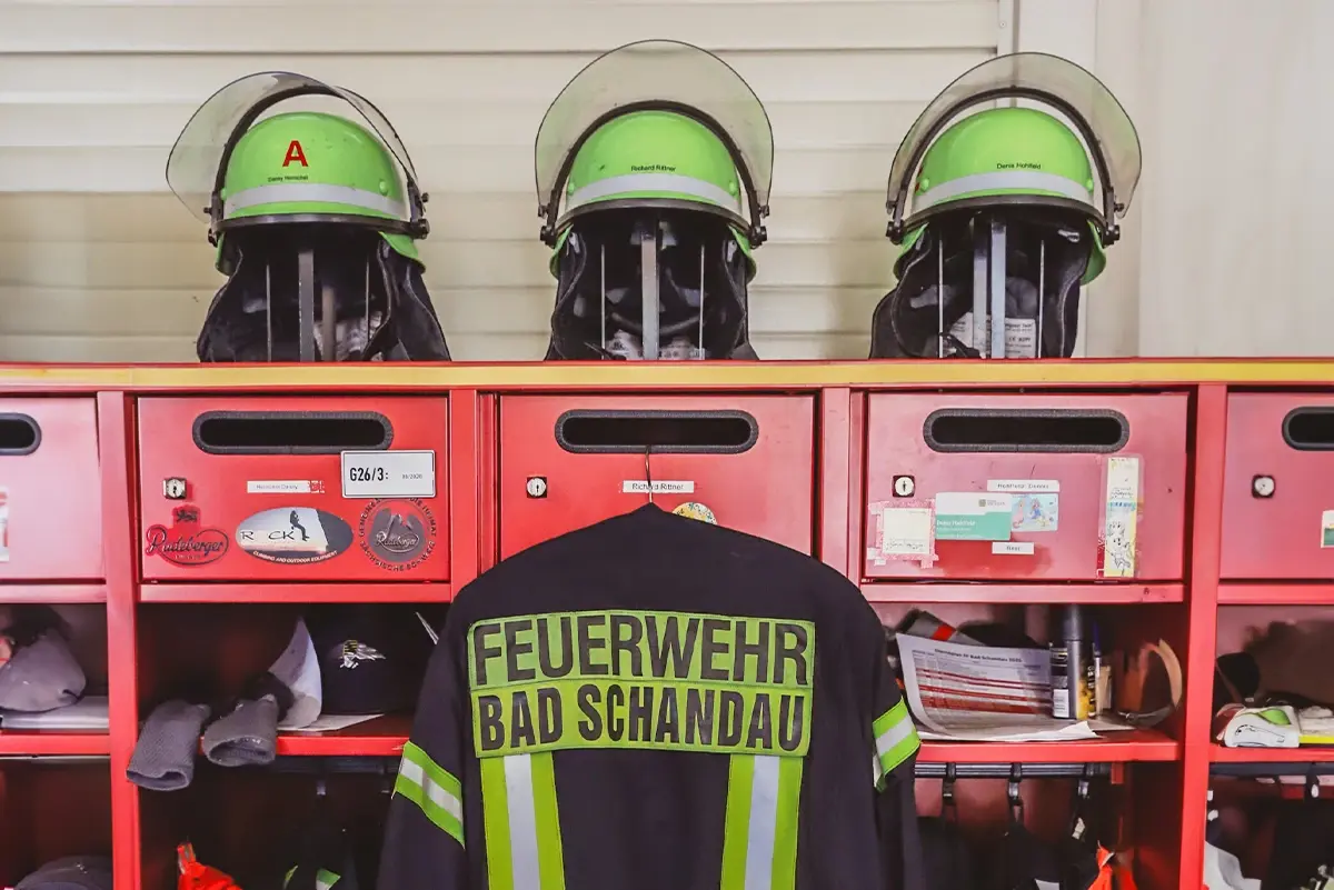 Drei Feuerwehrhelme ruhen auf roten Spinden, vor einem Einsatzmantel.