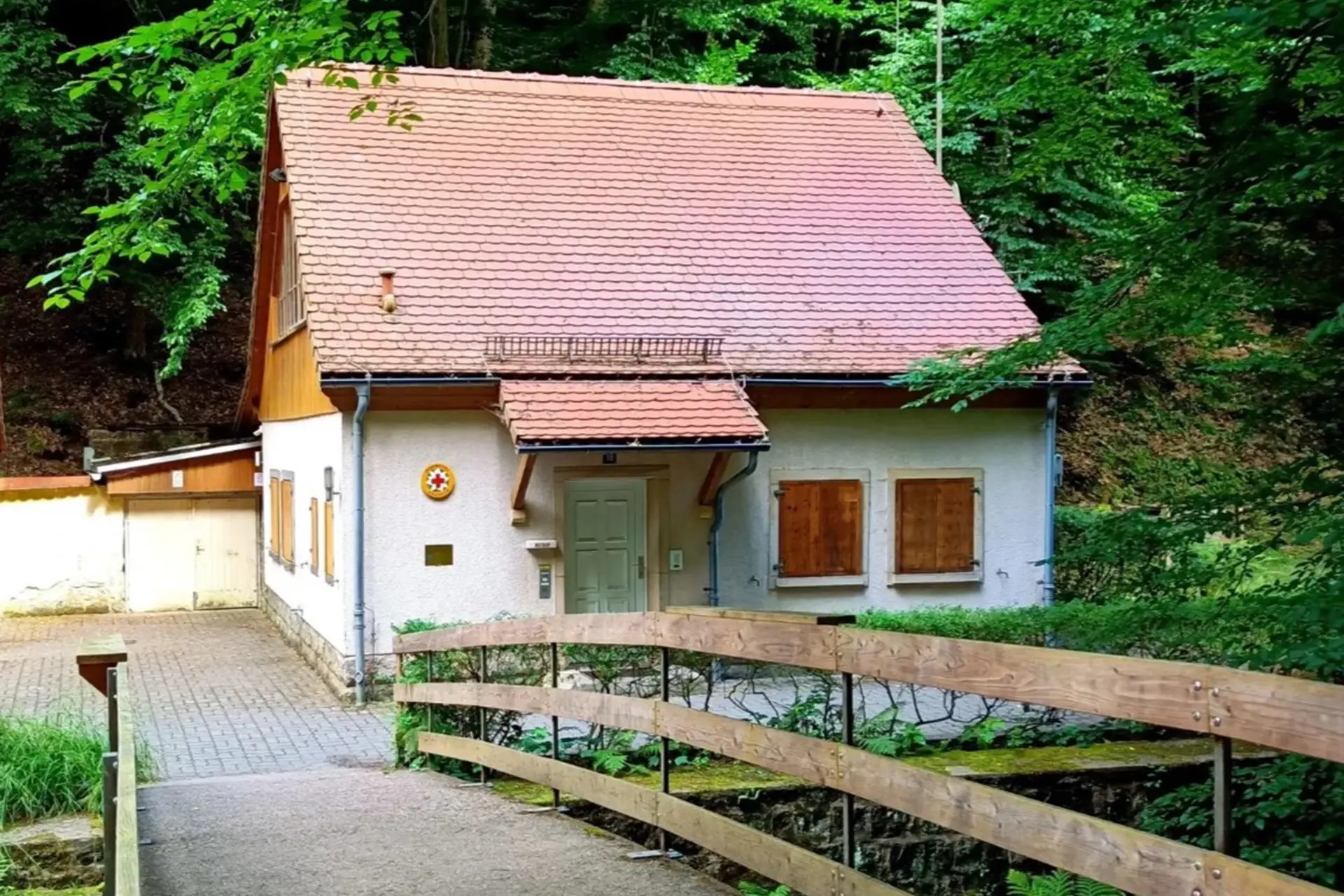 Ein kleines Haus steht umgeben von Bäumen mit einem rot gedeckten Dach und Holzzaun.