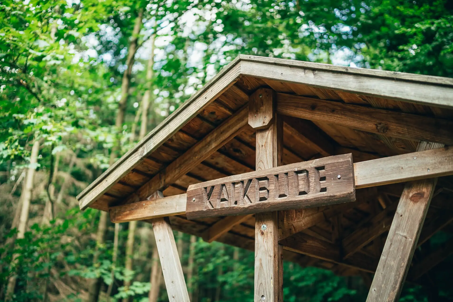Ein Holzdach in einem grünen Wald trägt das Schild „KAKBUDE“.