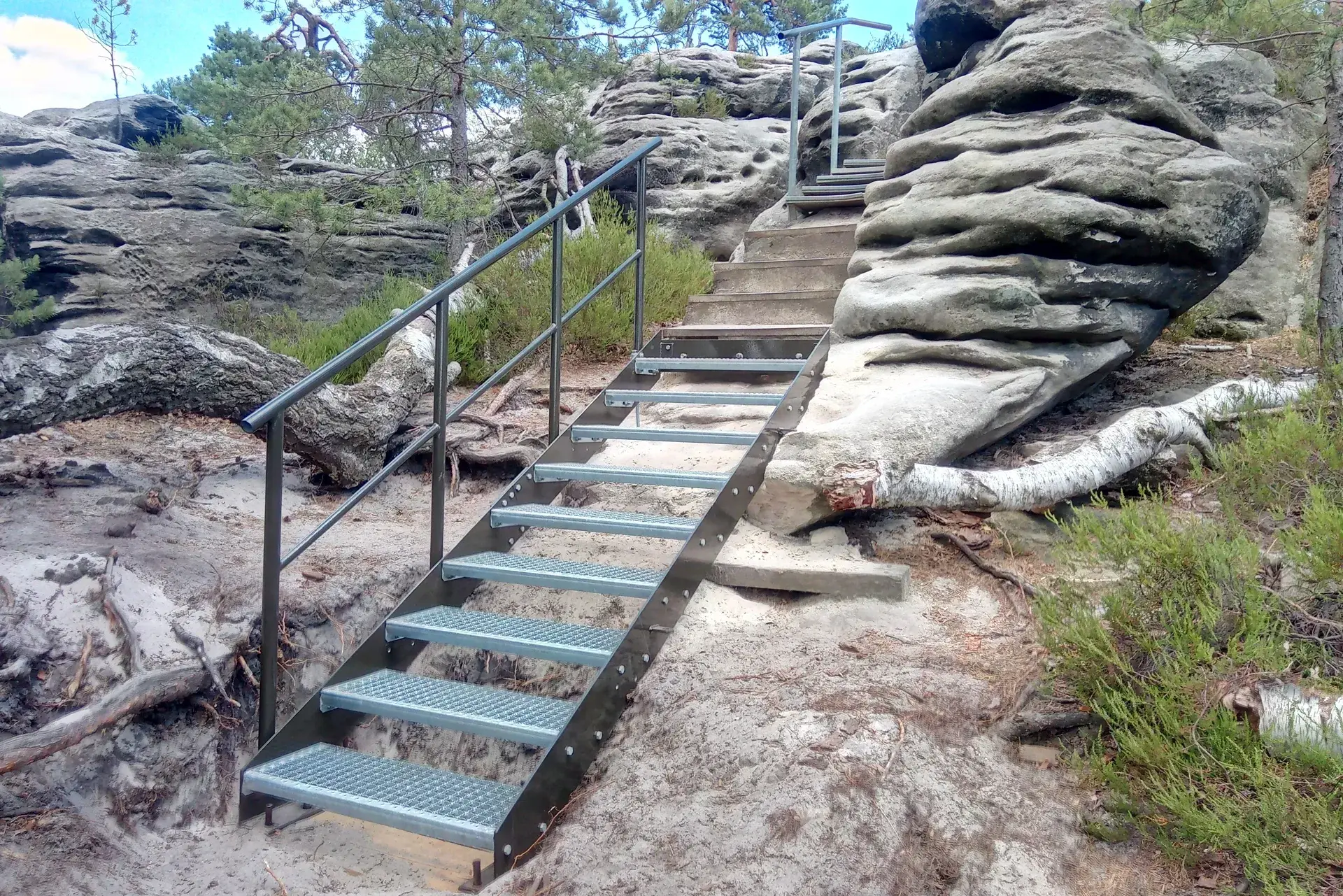 Metalltreppe führt auf einem felsigen Gelände hinauf, umgeben von Bäumen und einem umgestürzten Baumstamm.