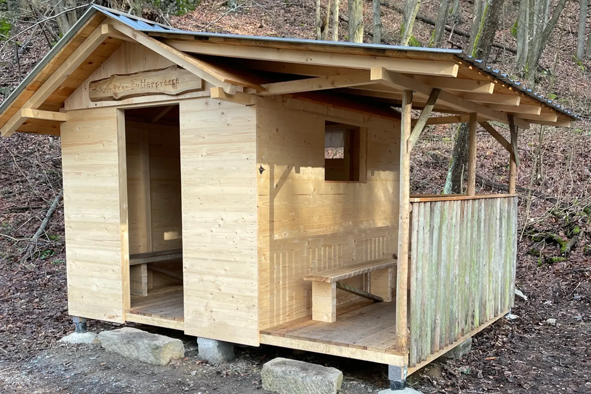 Kleine hölzerne Schutzhütte im Wald mit offenem Eingang und Sitzbank.