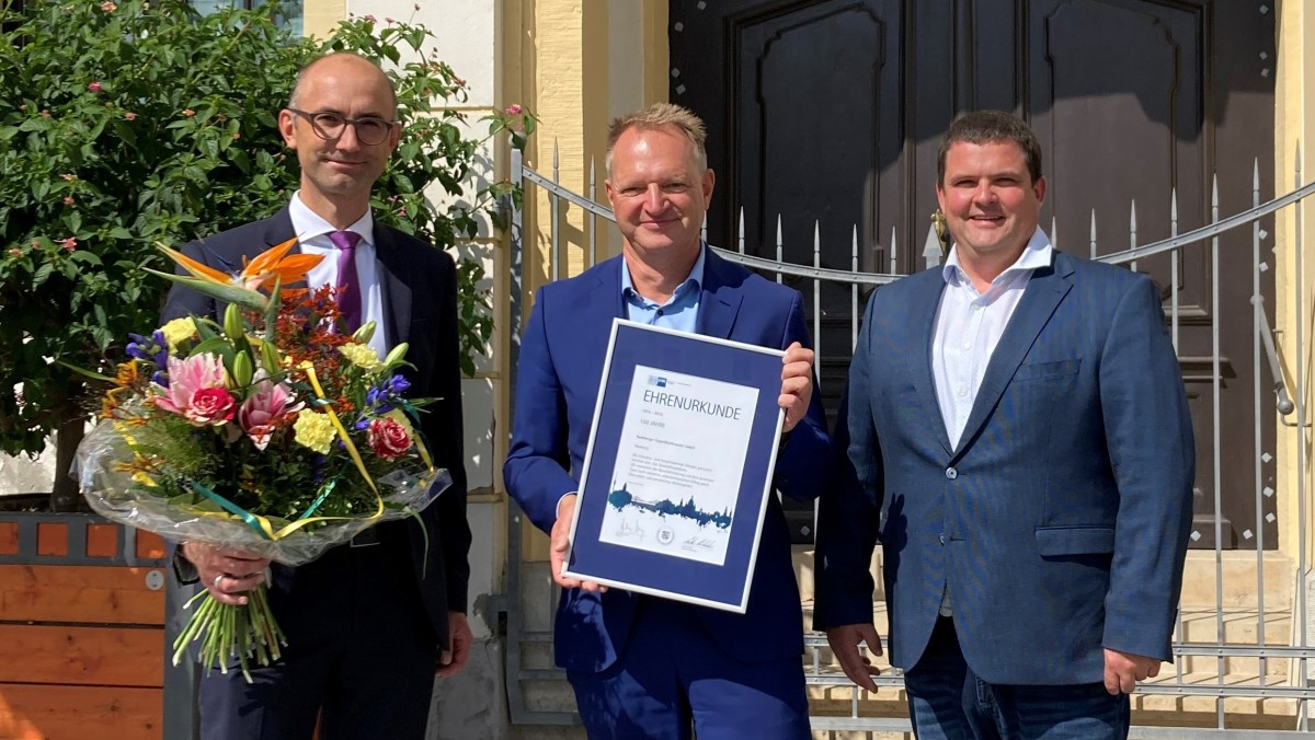 Drei Männer stehen vor einem Gebäude, einer hält eine Urkunde und Blumen.