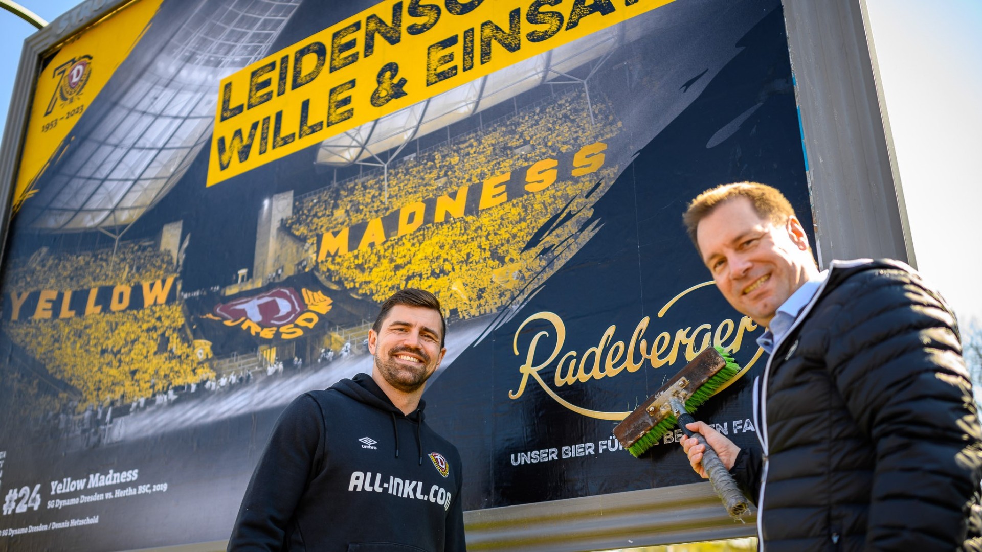 Zwei Männer stehen lächelnd vor einem großen Werbeplakat mit Stadionmotiv.