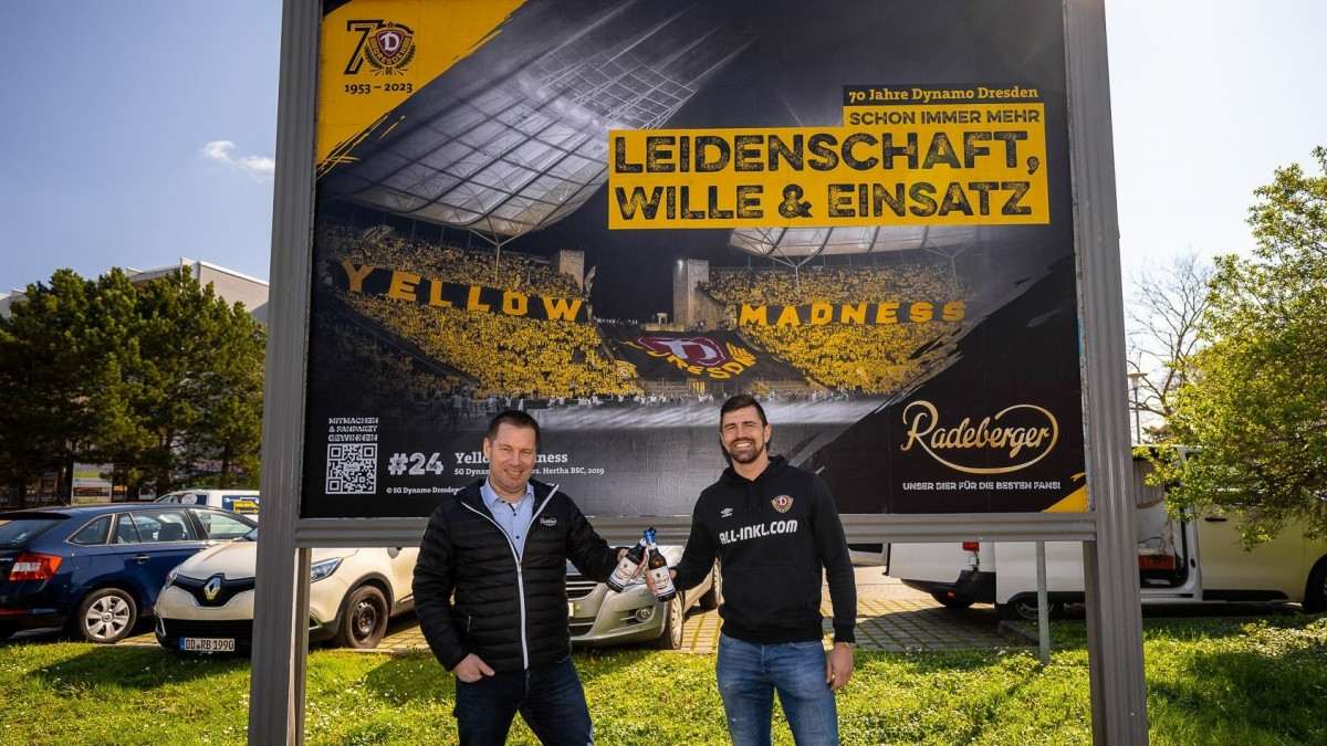 Zwei Männer stehen lächelnd vor einem großen Werbeplakat bei einer Sportveranstaltung.