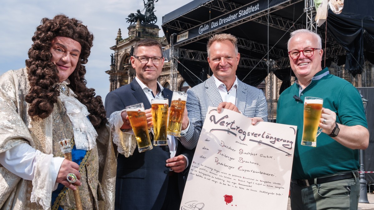 Vier Männer stehen lächelnd auf einer Bühne, einer in historischem Kostüm, drei mit Biergläsern und einem Plakat.