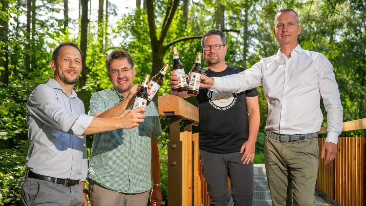 Vier Männer stehen im Freien und halten gemeinsam Bierflaschen hoch. Sie lächeln und sind lässig gekleidet.