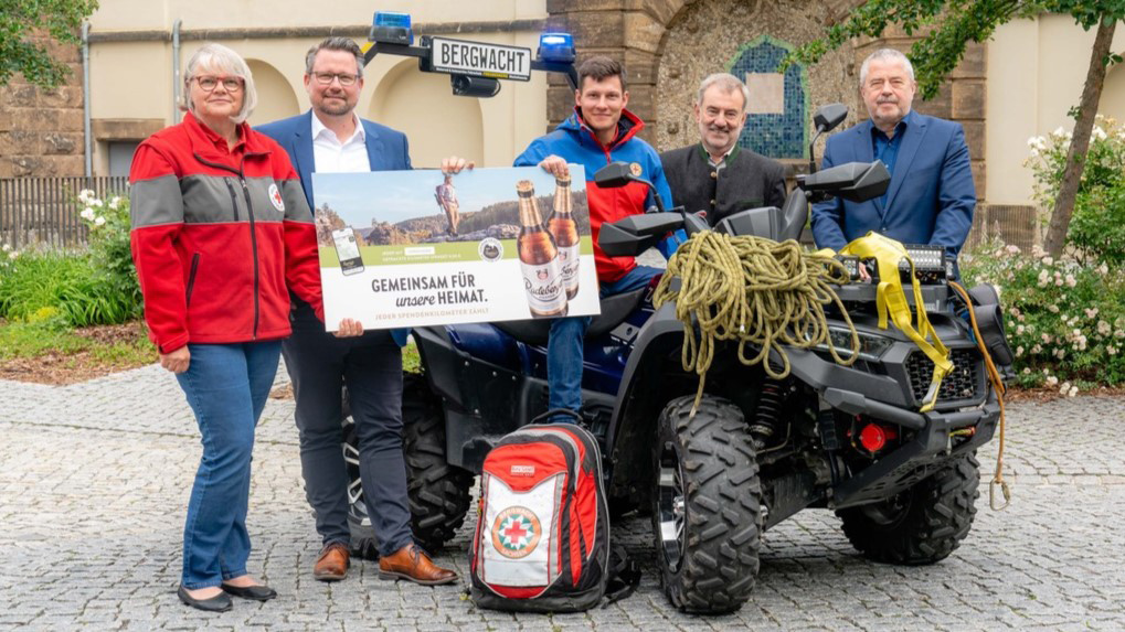 Fünf Personen stehen neben einem Quad vor einem historischen Gebäude. Sie halten eine Plakatwerbung und Rettungsausrüstung.