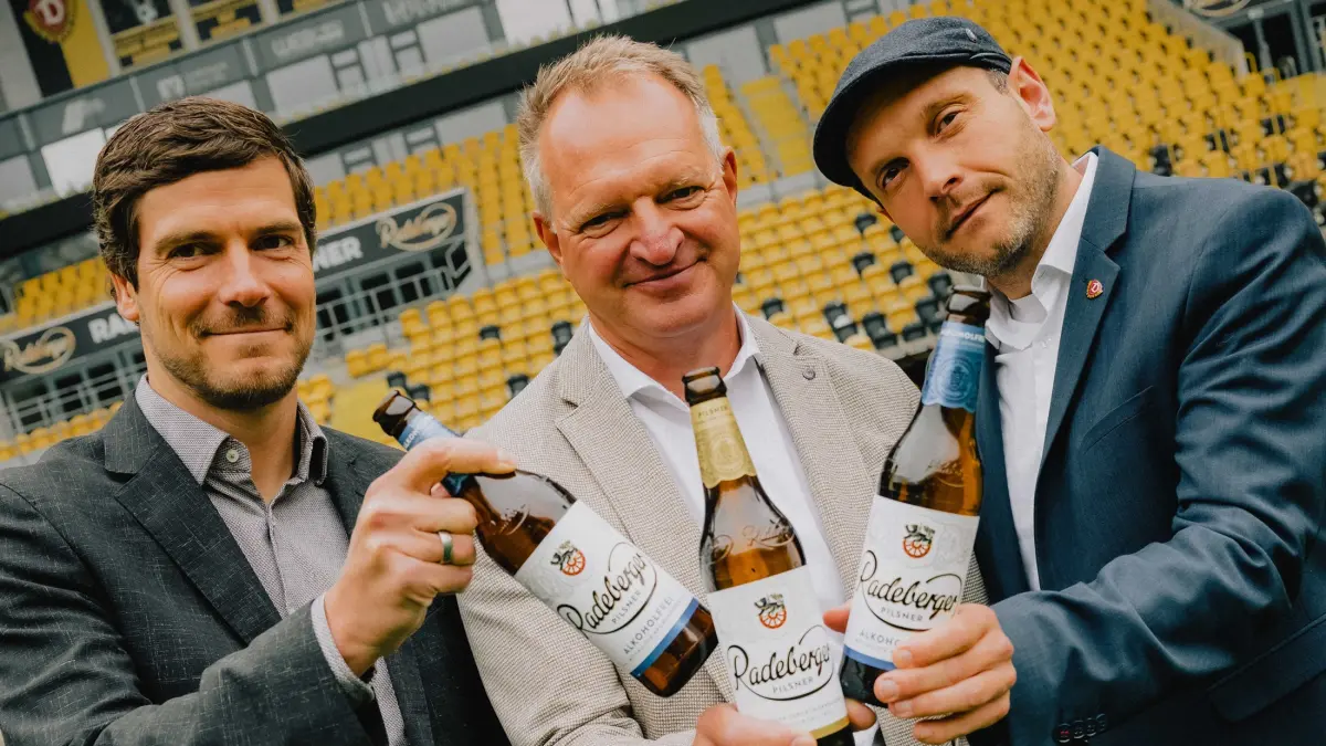 Drei Männer stehen in einem Stadion und halten Bierflaschen.