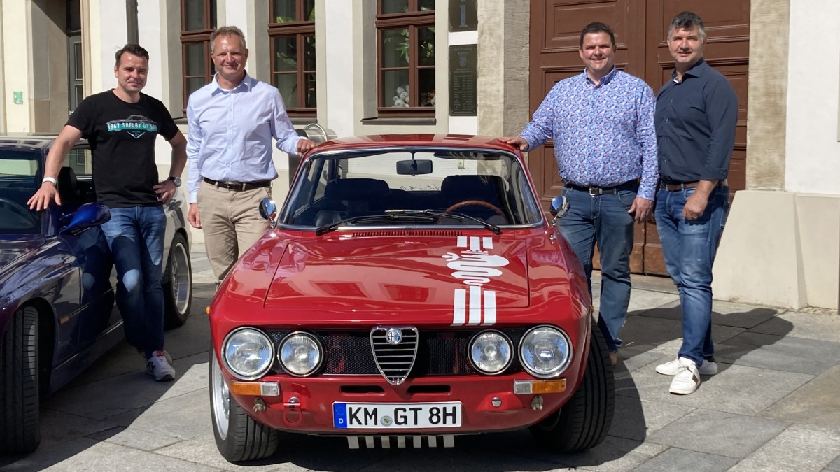 Vier Männer stehen neben einem klassischen roten Sportwagen vor einem Gebäude.
