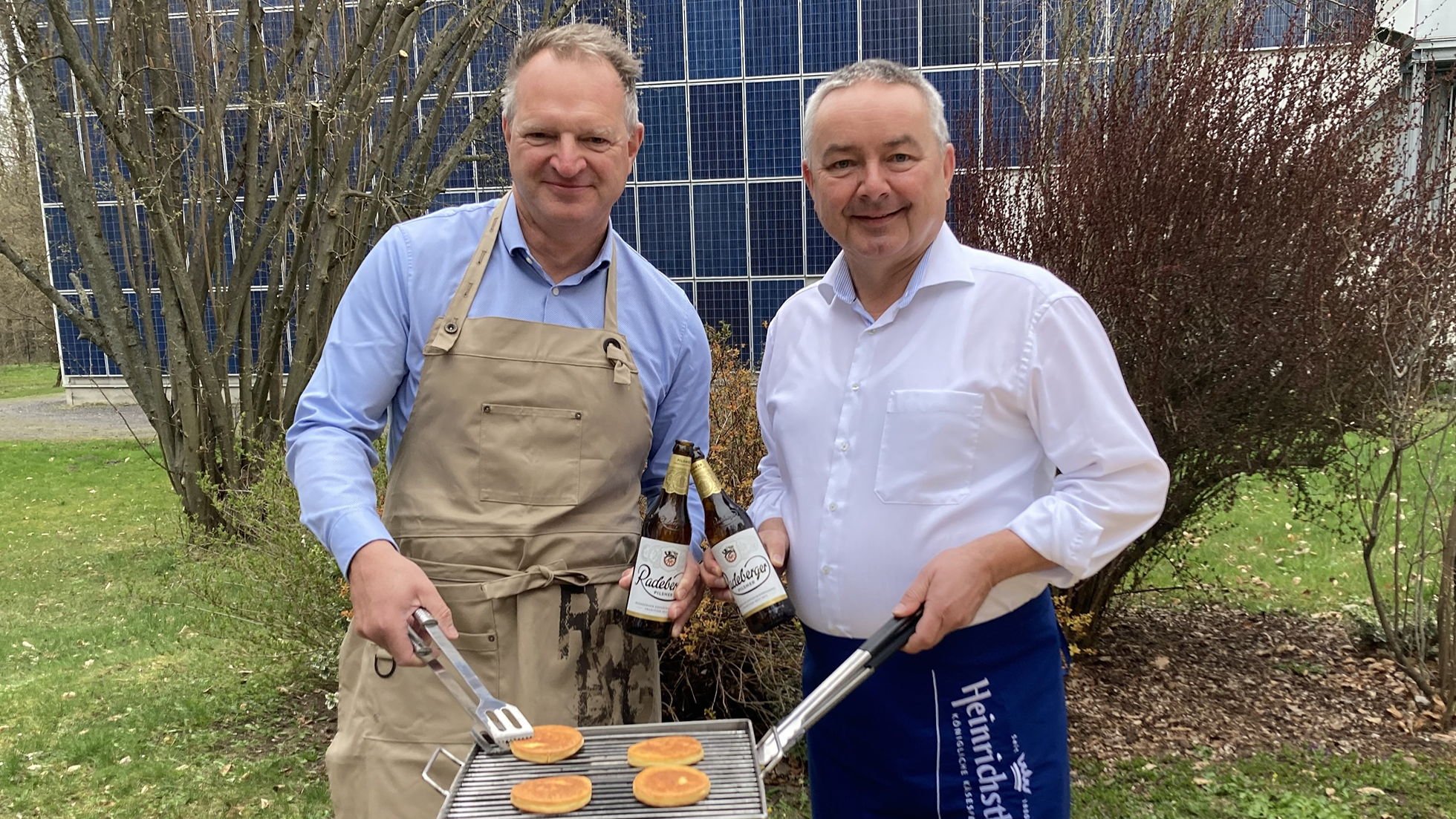 Zwei Männer stehen lächelnd neben einem Grill mit Pancakes; einer hält Bierflaschen.