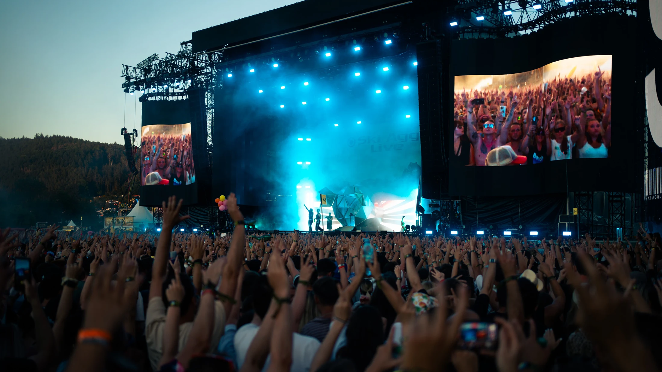 Menschenmenge mit erhobenen Armen vor einer großen Festivalbühne mit blauer Lichtshow. Im Hintergrund bewaldete Hügel.
